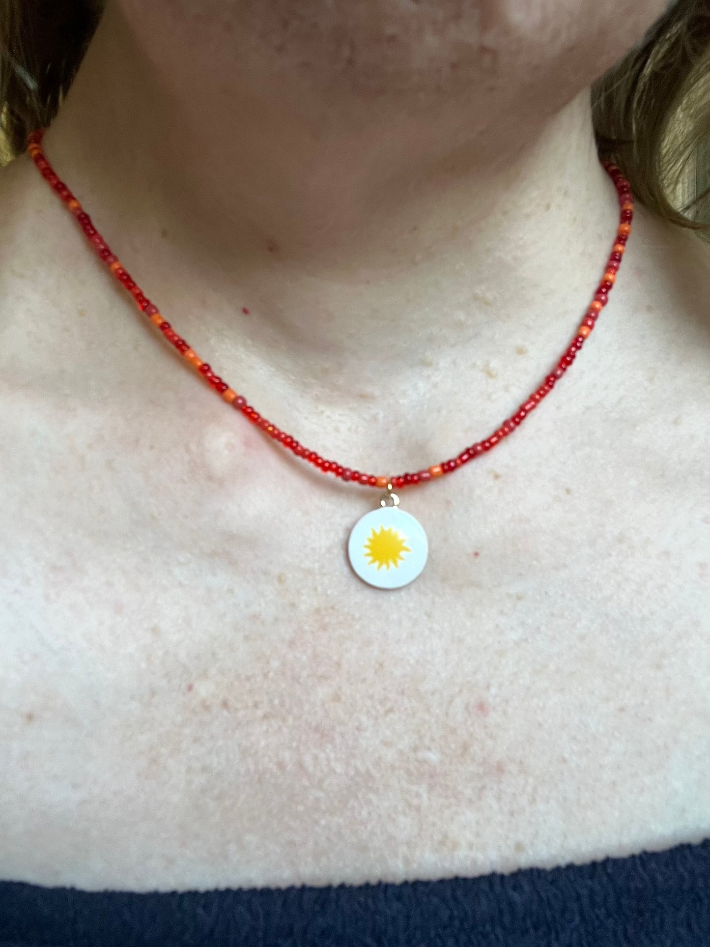 Collier fantaisie fait main.
Collier ras de cou en perles de rocaille rouge - orange.
Pendentif rond blanc orné d'un soleil.
Bijou fait main.