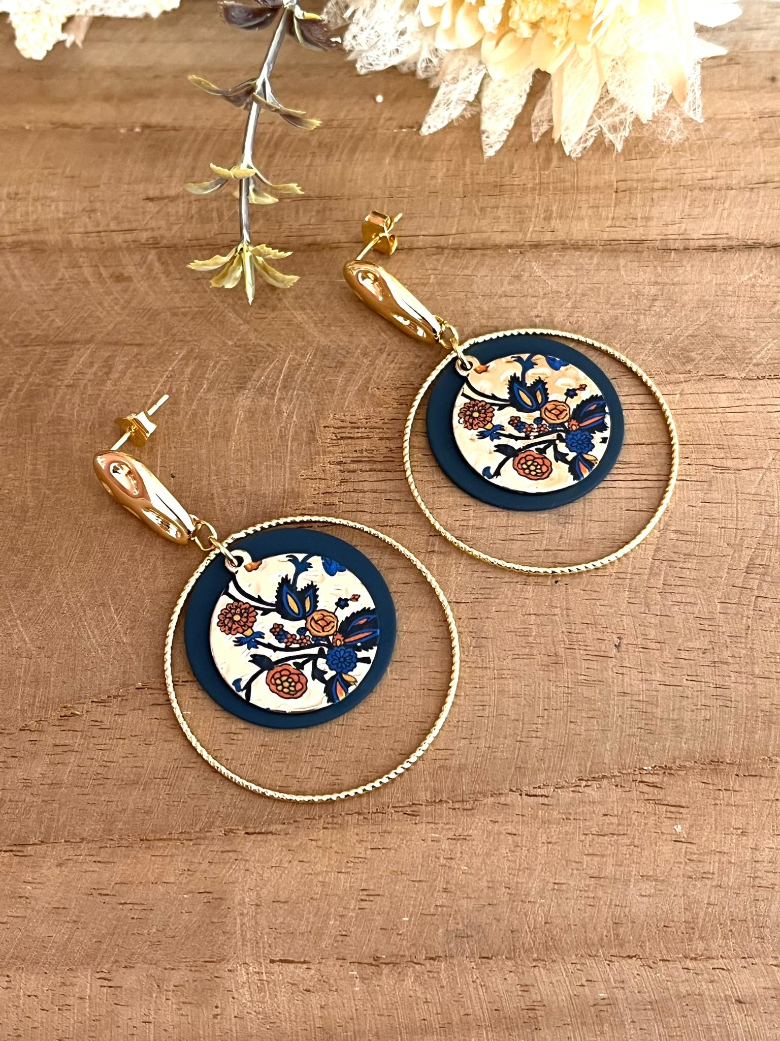 Boucles d'oreilles fantaisie fait main.
Clous d'oreilles martelés en laiton doré à l'or fin. Sequins ronds bleu marine, anneaux torsadés en laiton doré à l'or fin. Pendentif rond doré motif floral bleu et orange.
Bijou fantaisie fait main.