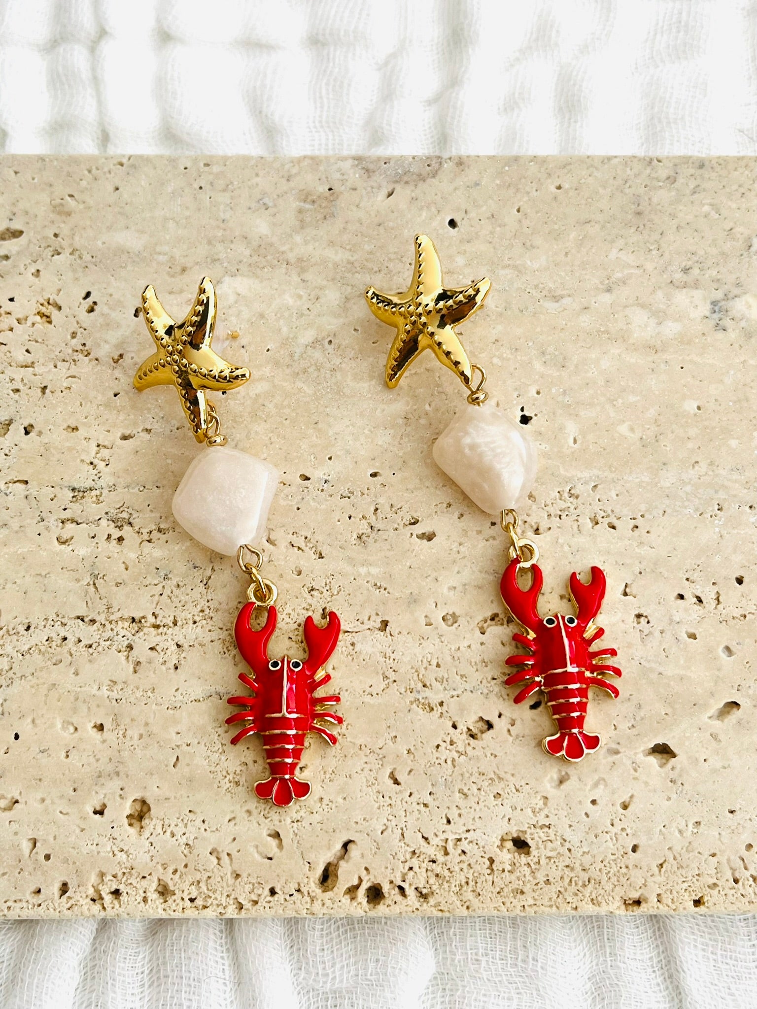 Boucles d'oreilles fantaisie fait main.
Clous d'oreilles étoile de mer laiton or 18k;
Perles connecteurs en acrylique blanc cassé,
breloques homard émaillé rouge.
Bijou fantaisie fait main.