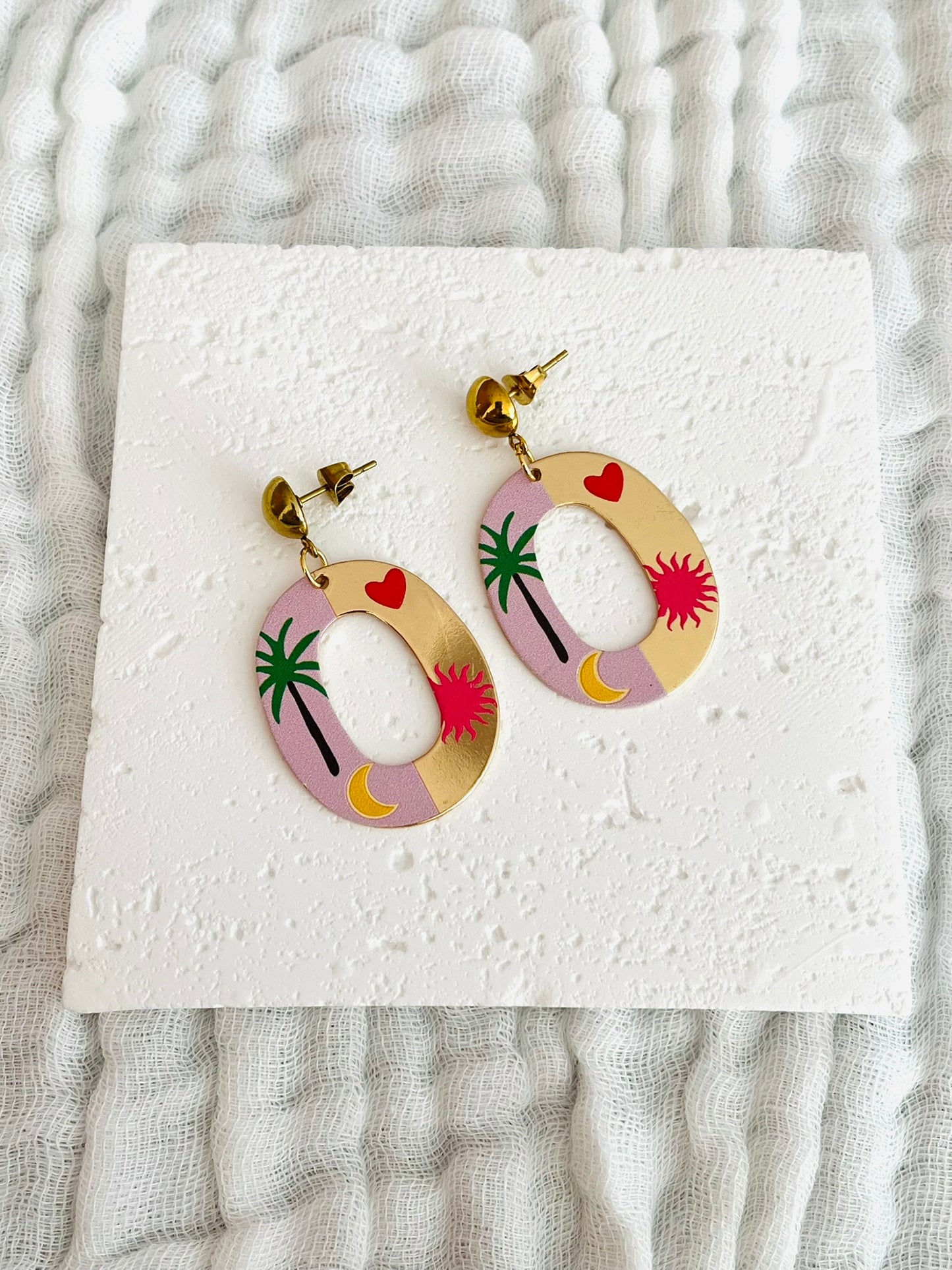 Boucles d'oreilles fantaisie fait main.
Clous d'oreilles en acier inox doré.
Pendentif ovale doré et rose avec motif palmier, soleil, lune, coeur.
Bijou fait main.
