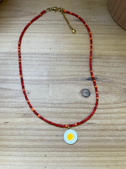 Collier fantaisie fait main.
Collier ras de cou en perles de rocaille rouge - orange.
Pendentif rond blanc orné d'un soleil.
Bijou fait main.