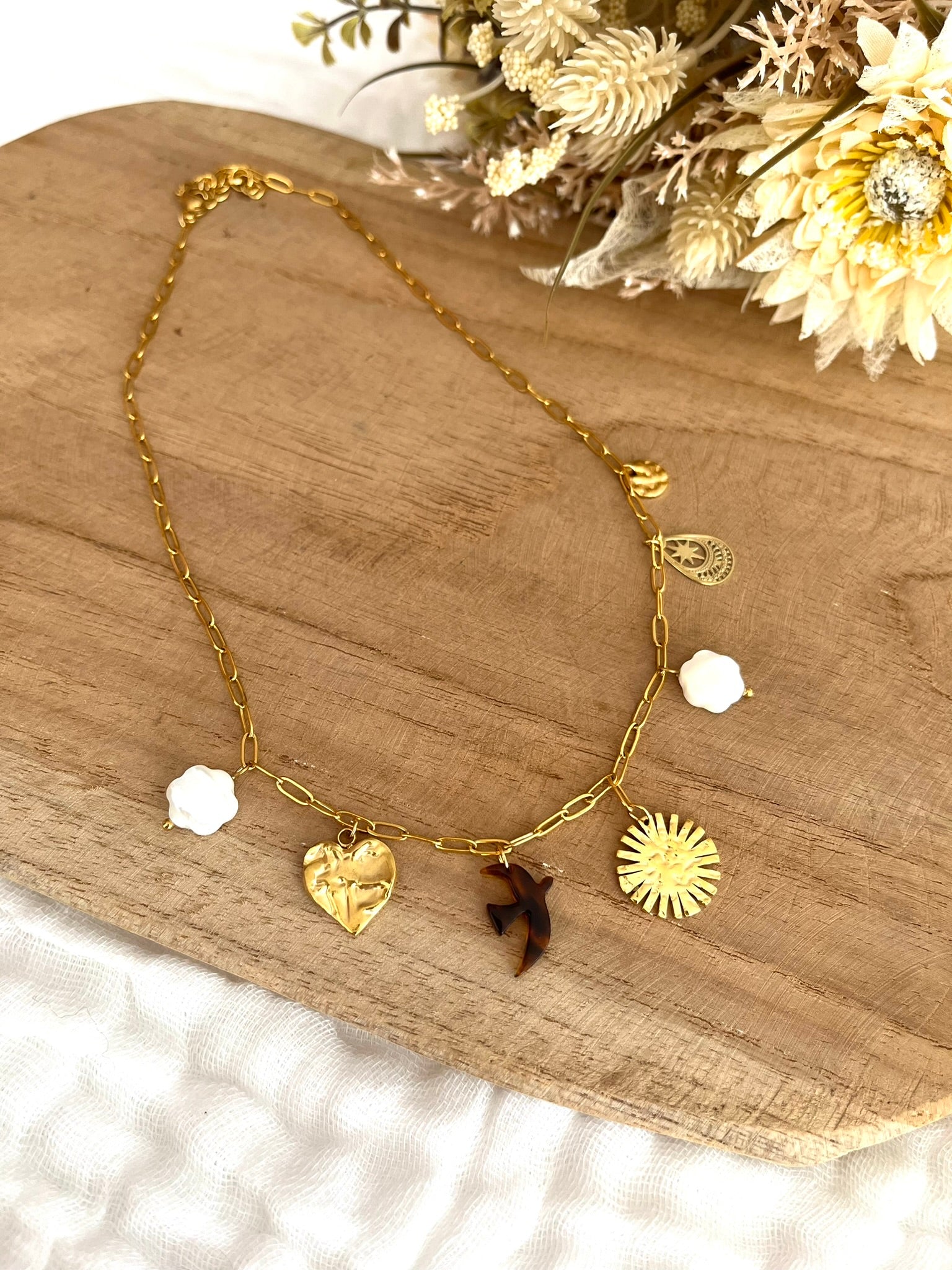 Collier fantaisie fait main.
Collier court en acier inoxydable doré avec 7 charms: fleurs en nacre, soleil, coeur, oiseau en résine, goutte ethnique, sequin martelé.
Bijou fantaisie fait main