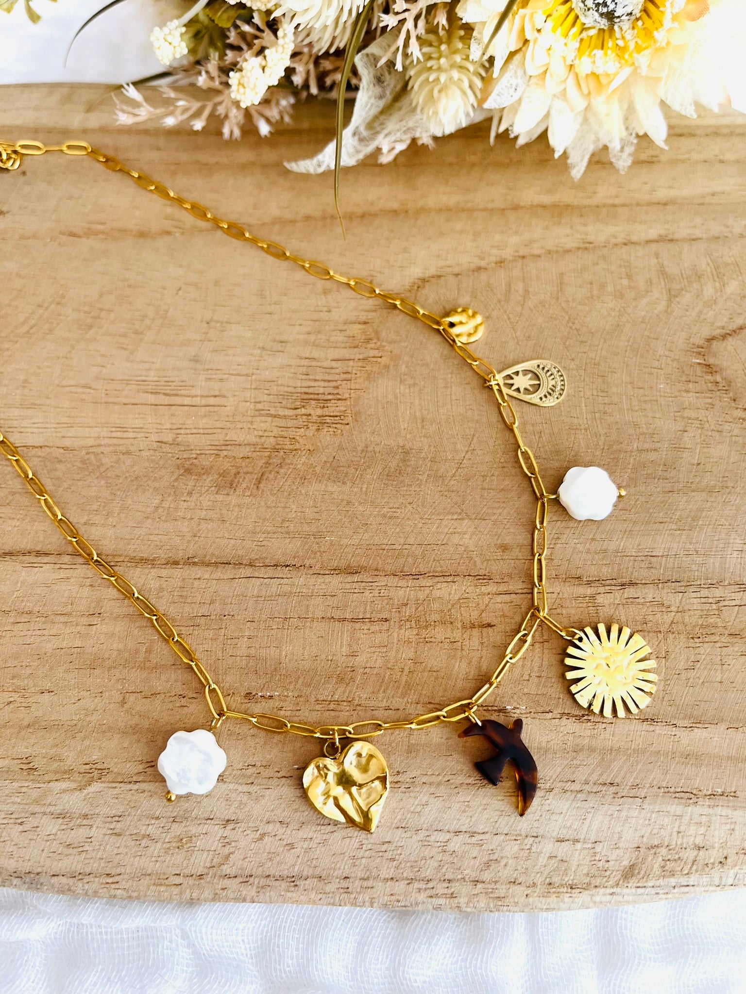 Collier fantaisie fait main.
Collier court en acier inoxydable doré avec 7 charms: fleurs en nacre, soleil, coeur, oiseau en résine, goutte ethnique, sequin martelé.
Bijou fantaisie fait main