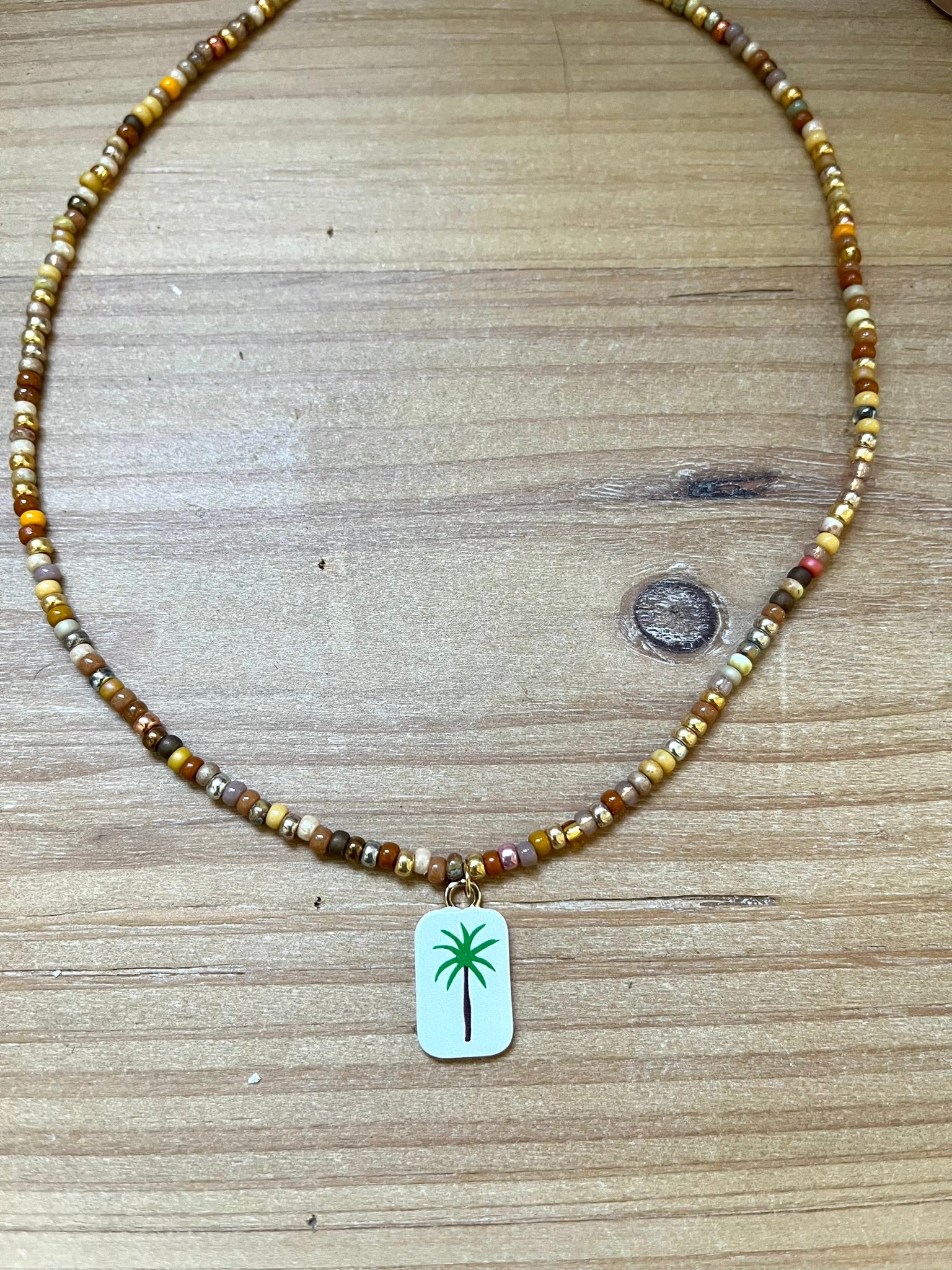 Collier fantaisie fait main.
Collier ras de cou en perles de rocaille dans les tons chauds de doré, marron, terre.
Pendentif rectangulaire blanc orné d'un palmier.
Bijou fait main.