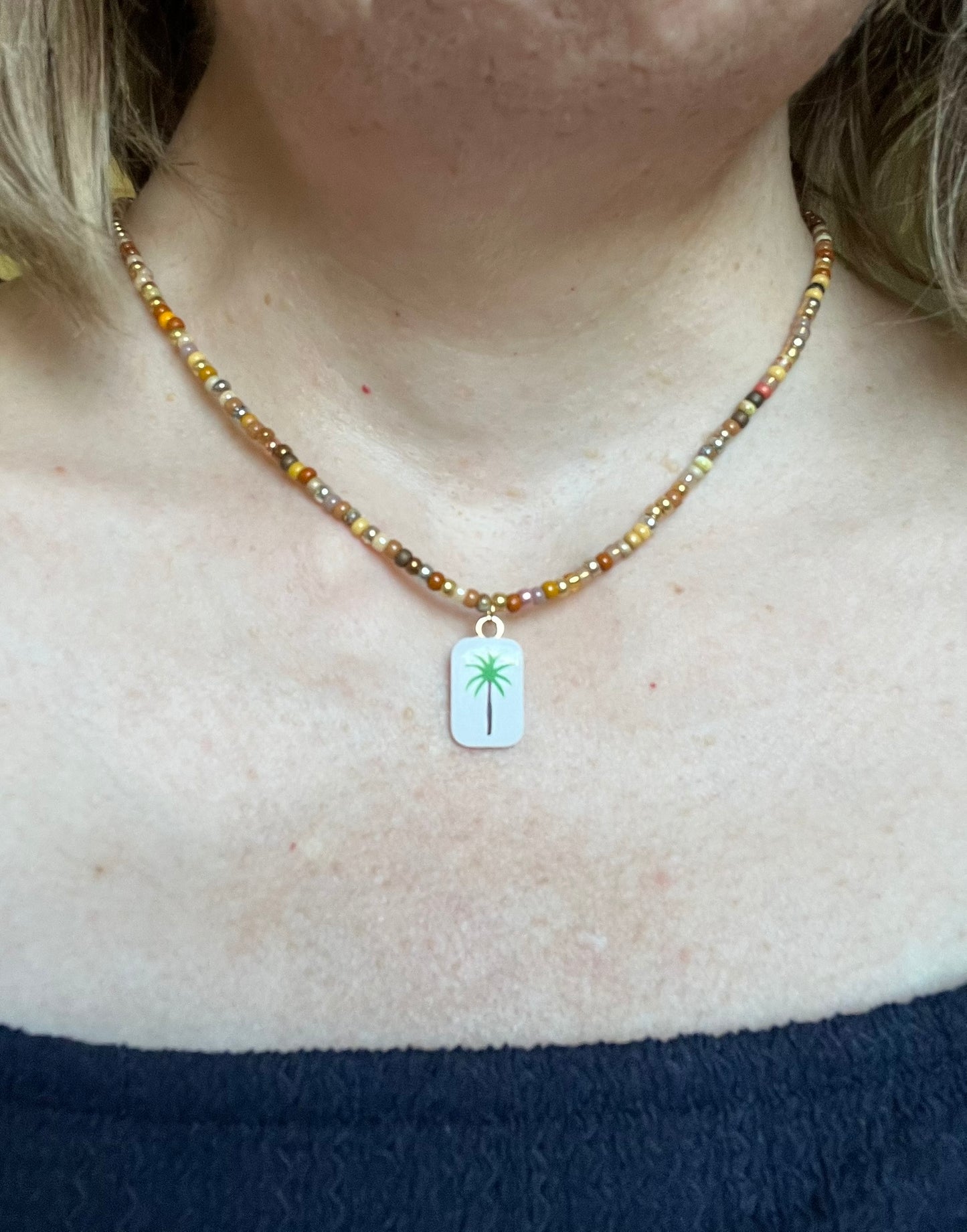 Collier fantaisie fait main.
Collier ras de cou en perles de rocaille dans les tons chauds de doré, marron, terre.
Pendentif rectangulaire blanc orné d'un palmier.
Bijou fait main.
