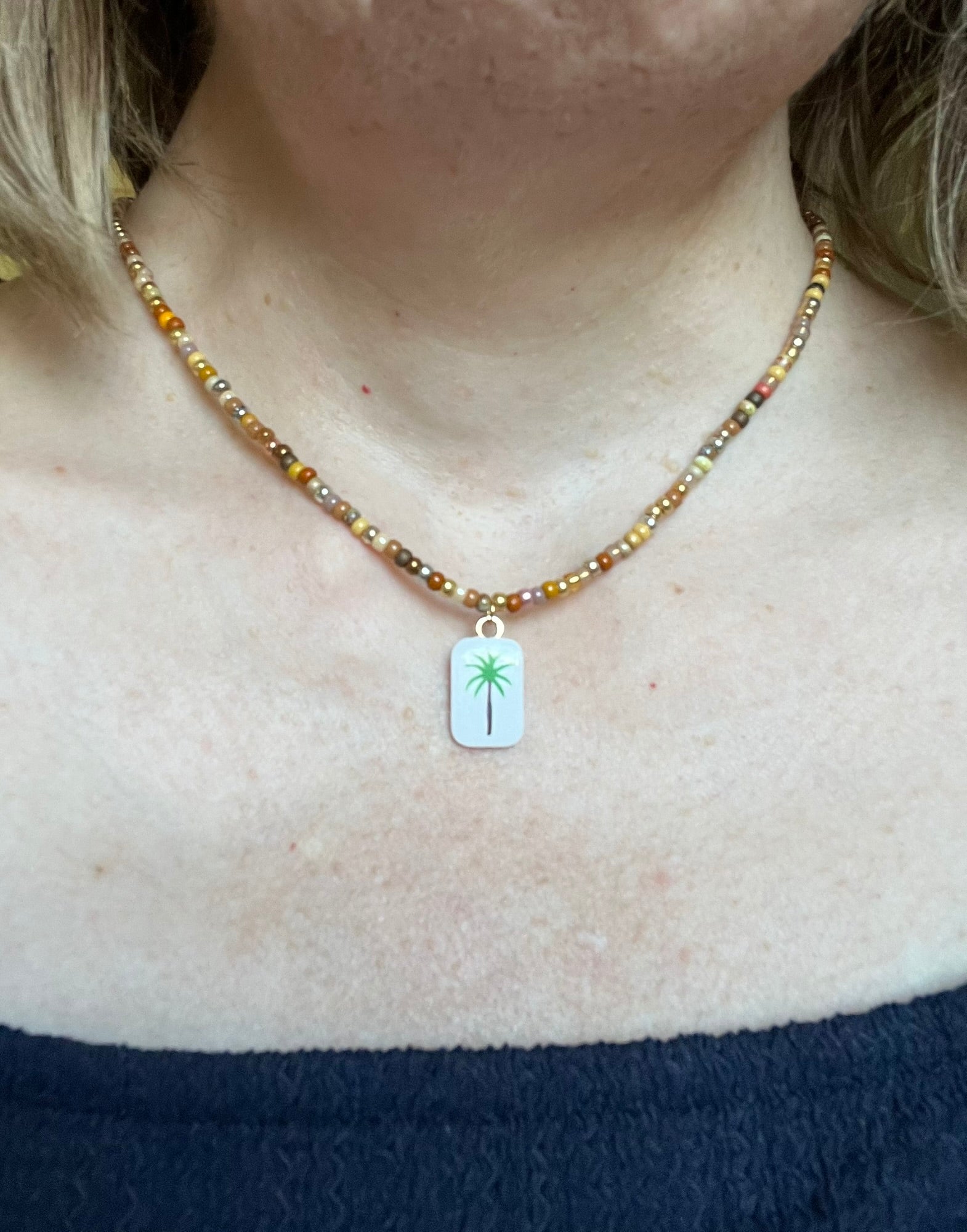 Collier fantaisie fait main.
Collier ras de cou en perles de rocaille dans les tons chauds de doré, marron, terre.
Pendentif rectangulaire blanc orné d'un palmier.
Bijou fait main.