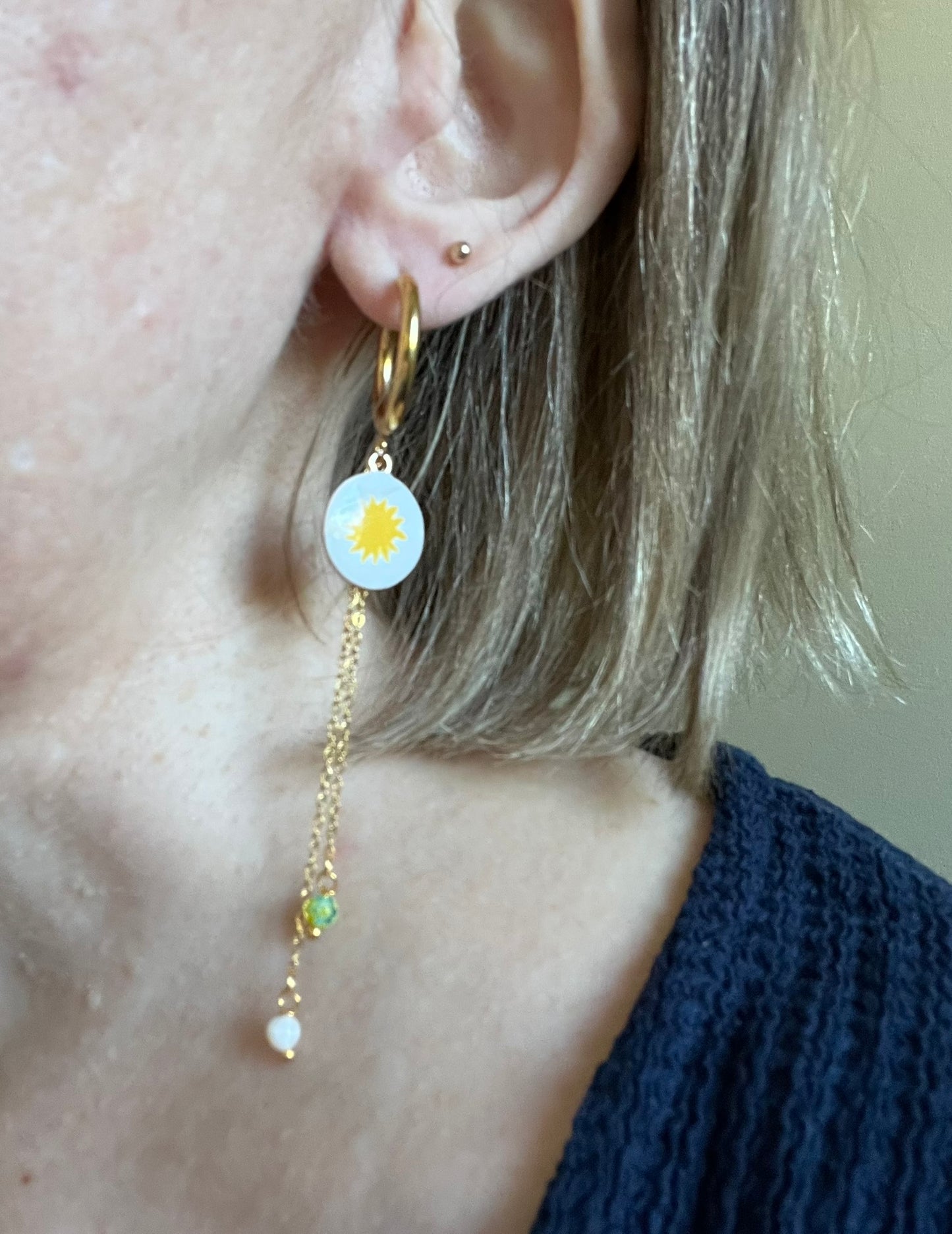 Boucles d'oreilles fantaisie fait main.
Créoles en acier doré.
Pendentif blanc blanc orné d'un soleil jaune.
2 Chainettes de longueur asymétrique réhaussées de perles millefiori dans les tons jaune et blanc.
Bijou fait main
